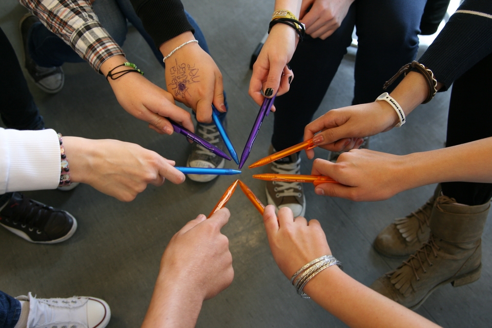 Schülerinnen und Schüler halten Spitzen ihrer bunten Kugelschreiber im Kreis zusammen